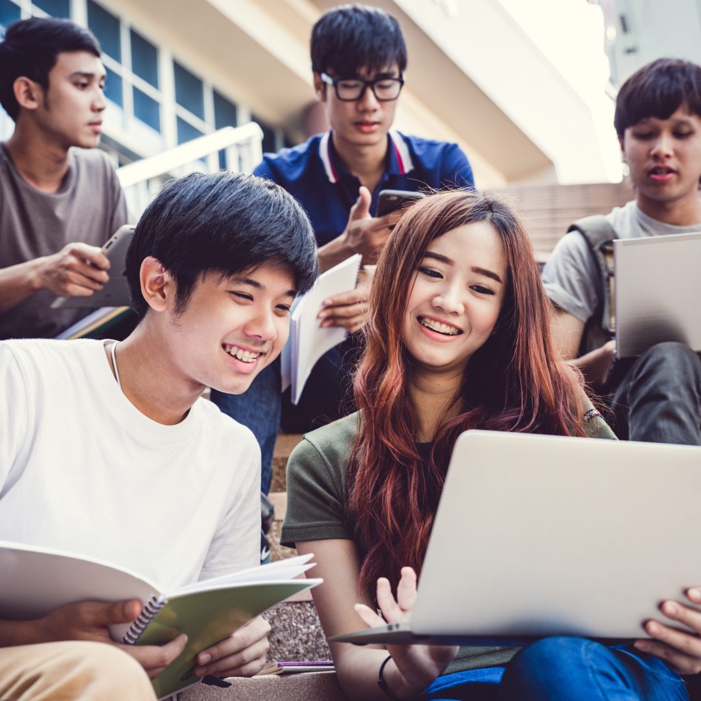 Group of JC Economics students studying together outdoors, discussing notes and reviewing tuition materials on a laptop